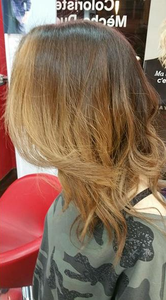 Femme aux cheveux bruns, avec un ombré blond, coiffés en ondulations. Elle porte une chemise camouflage.