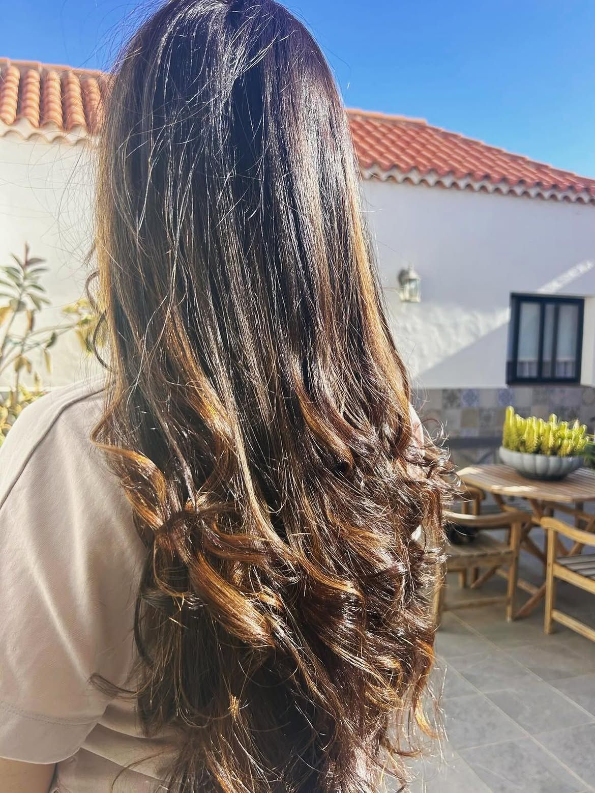 Cabello largo, oscuro y ondulado bajo el sol, visto desde atrás frente a un patio de estuco blanco con tejas de terracota.