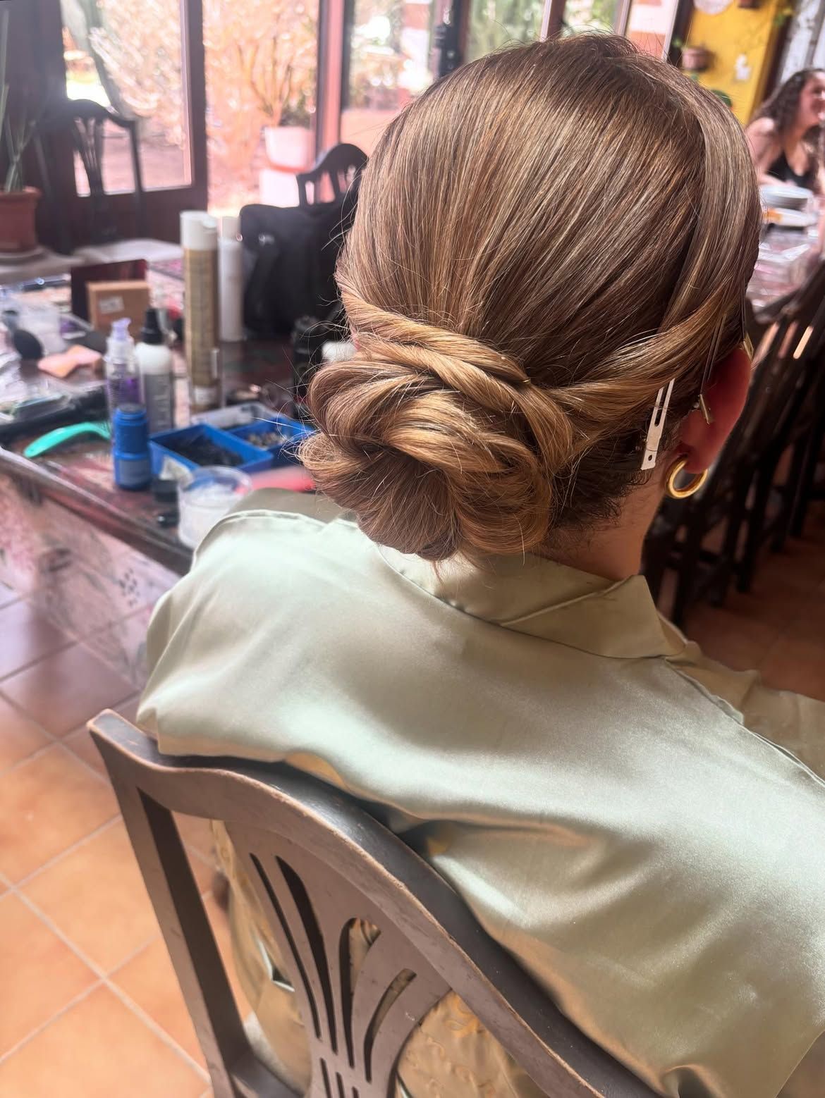 Persona con un moño bajo trenzado sentada en una silla de peluquería, vista desde atrás, con un espejo y una mesa delante.