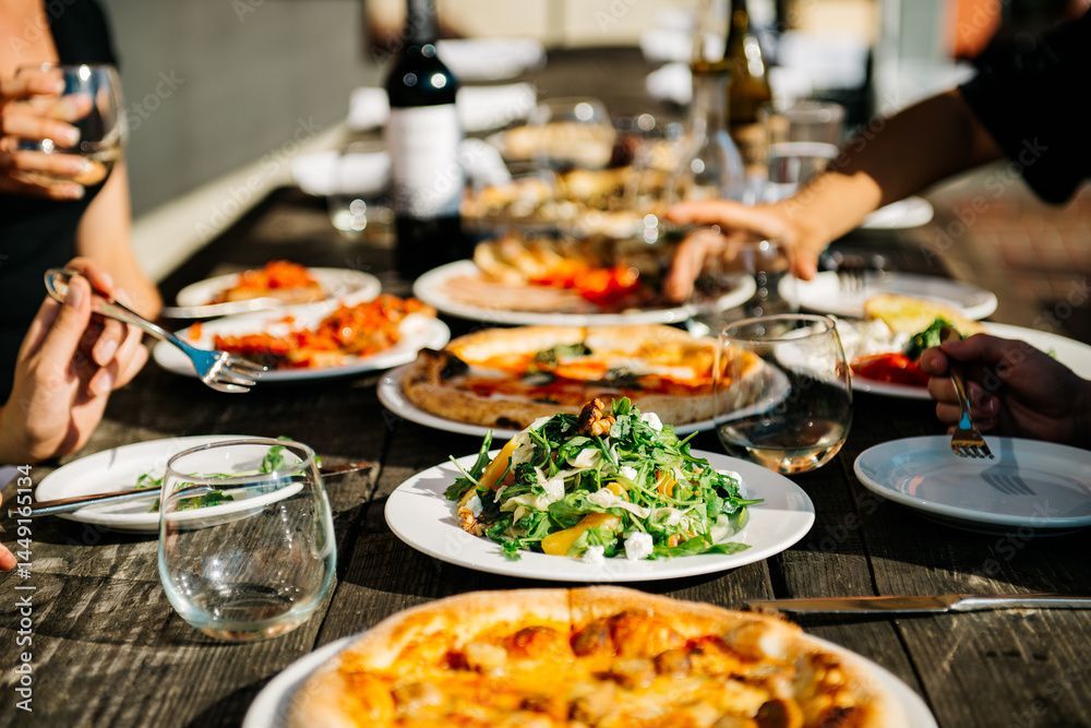 Menschen greifen nach Essen an einem rustikalen Holztisch: Pizza, Salat und Wein.