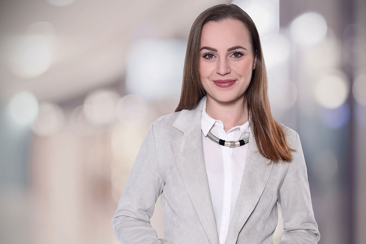 Eine Frau in einer grauen Jacke und einem weißen Hemd steht vor einem unscharfen Hintergrund.