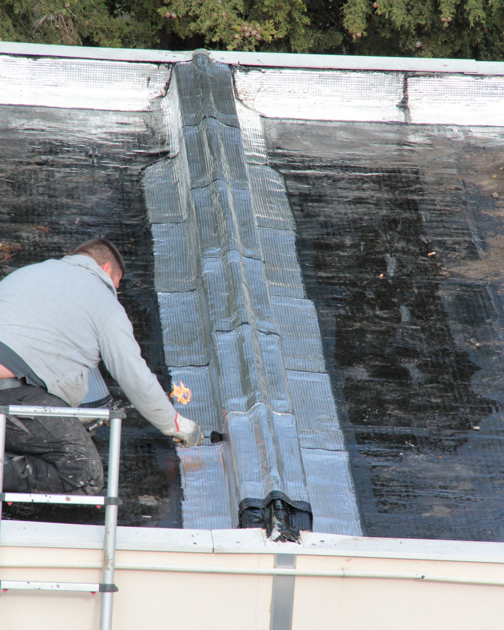 Un couvreur applique du mastic d'étanchéité sur un toit plat à l'aide d'une échelle.