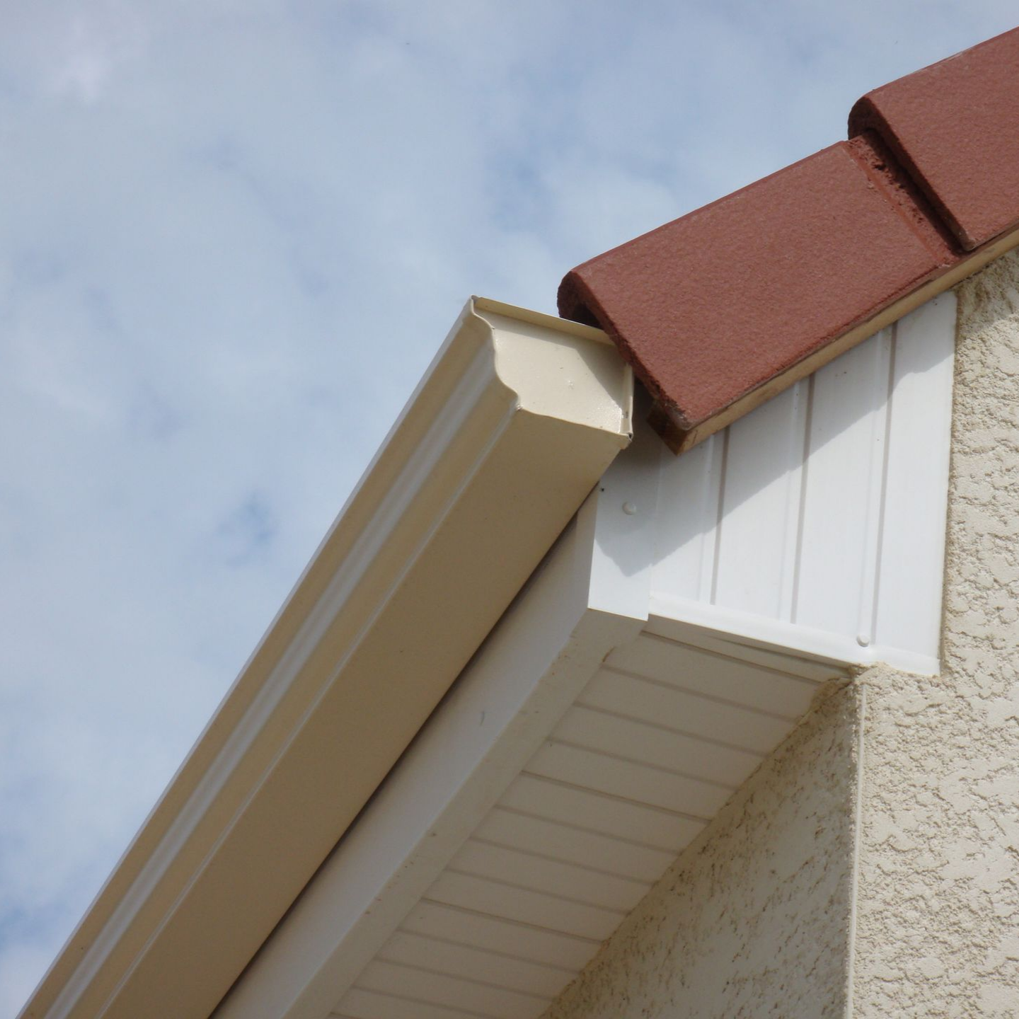 Une gouttière beige longe un toit de tuiles couleur terre cuite sur fond de ciel nuageux.