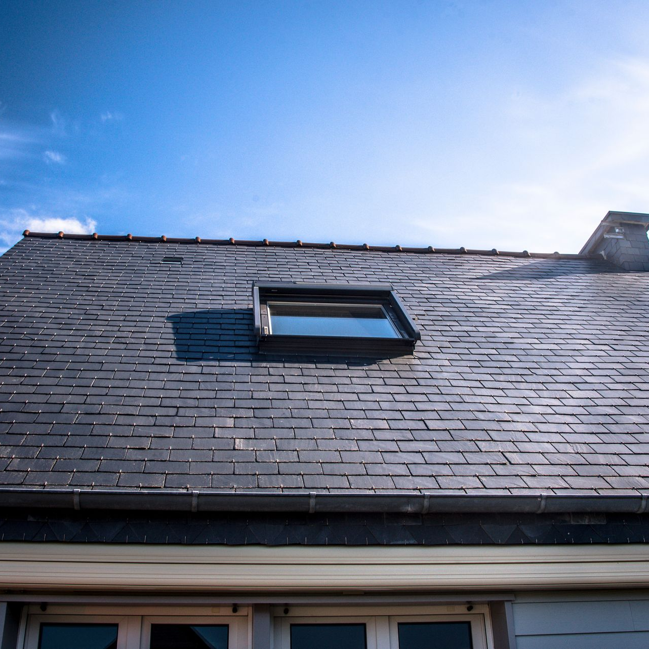 Toit en ardoise avec une fenêtre de toit sur fond de ciel bleu.