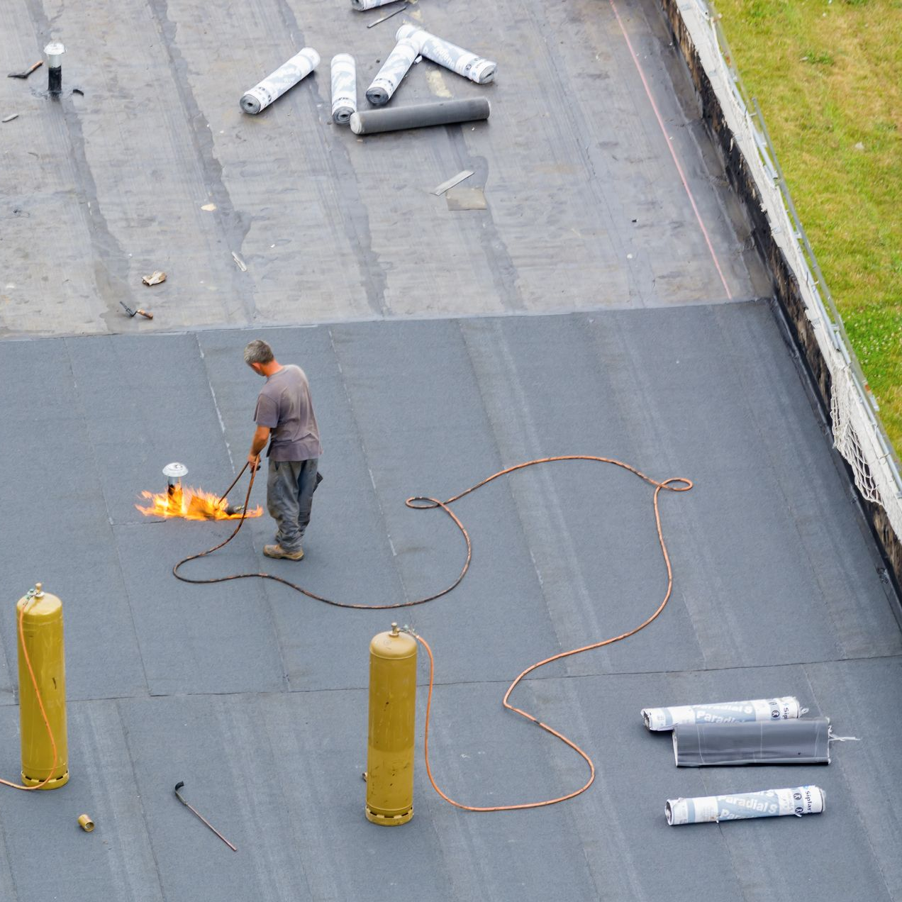 Un couvreur utilise un chalumeau sur un toit plat près de rouleaux d'étanchéité.