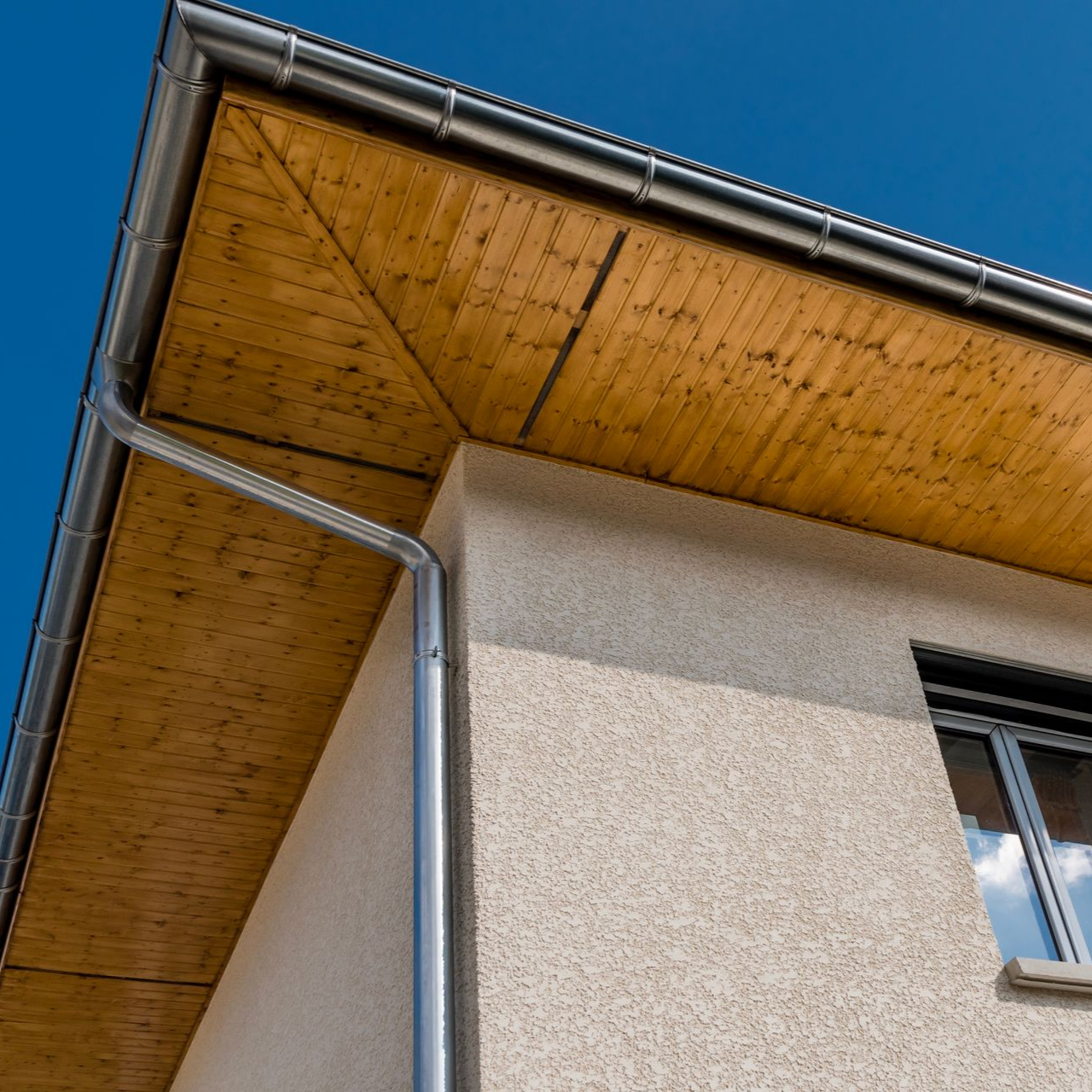 Coin d'une maison avec sous-face en bois et gouttières argentées sur fond de ciel bleu.