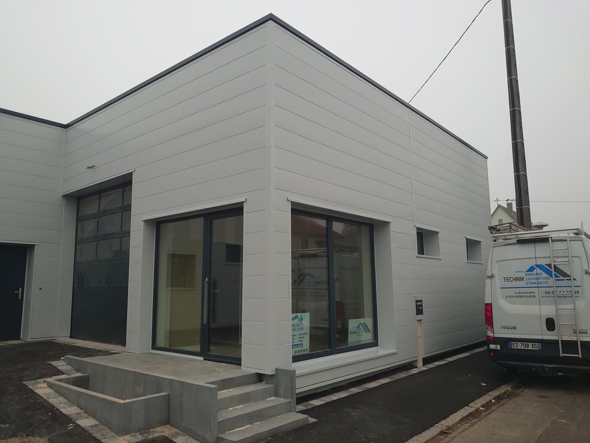 Bâtiment blanc moderne aux portes et fenêtres sombres, avec des marches menant à l'entrée.