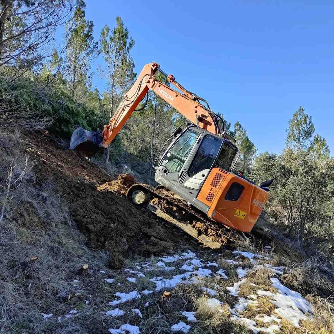 Excavadora naranja en una ladera embarrada, despejando maleza. Cielo azul brillante, paisaje invernal.