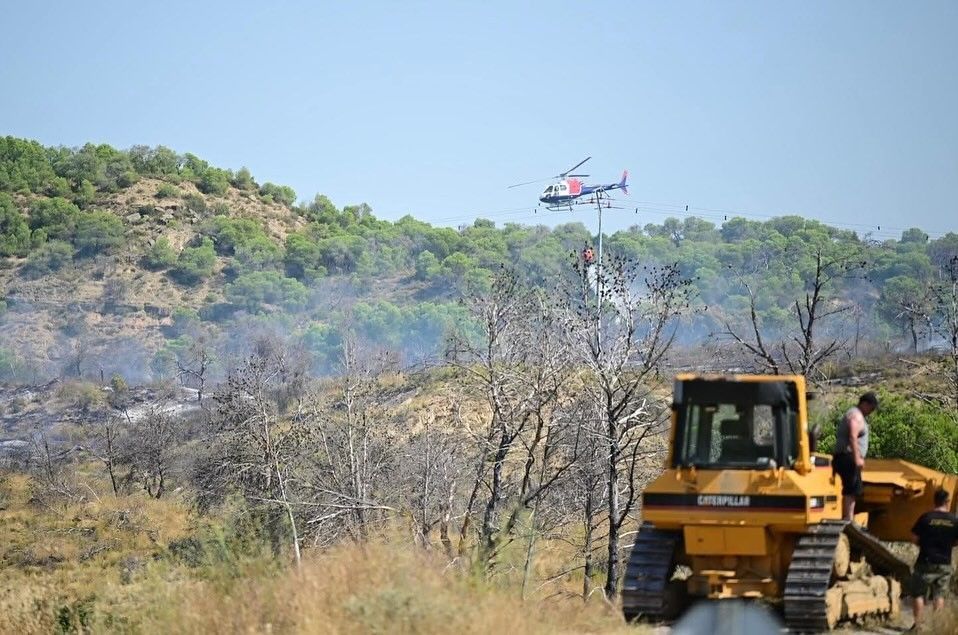 Helicóptero lanzando agua sobre un incendio forestal. Bulldozer en el suelo, con humo elevándose.