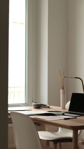Un escritorio blanco minimalista con una taza beige, un cuaderno, un lápiz, un cuenco pequeño y un jarrón alto con hierba de la pampa junto a una silla.