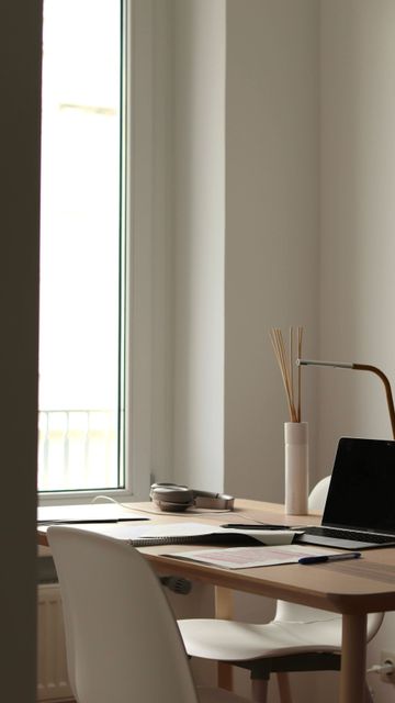 Un escritorio blanco minimalista con una taza beige, un cuaderno, un lápiz, un cuenco pequeño y un jarrón alto con hierba de la pampa junto a una silla.
