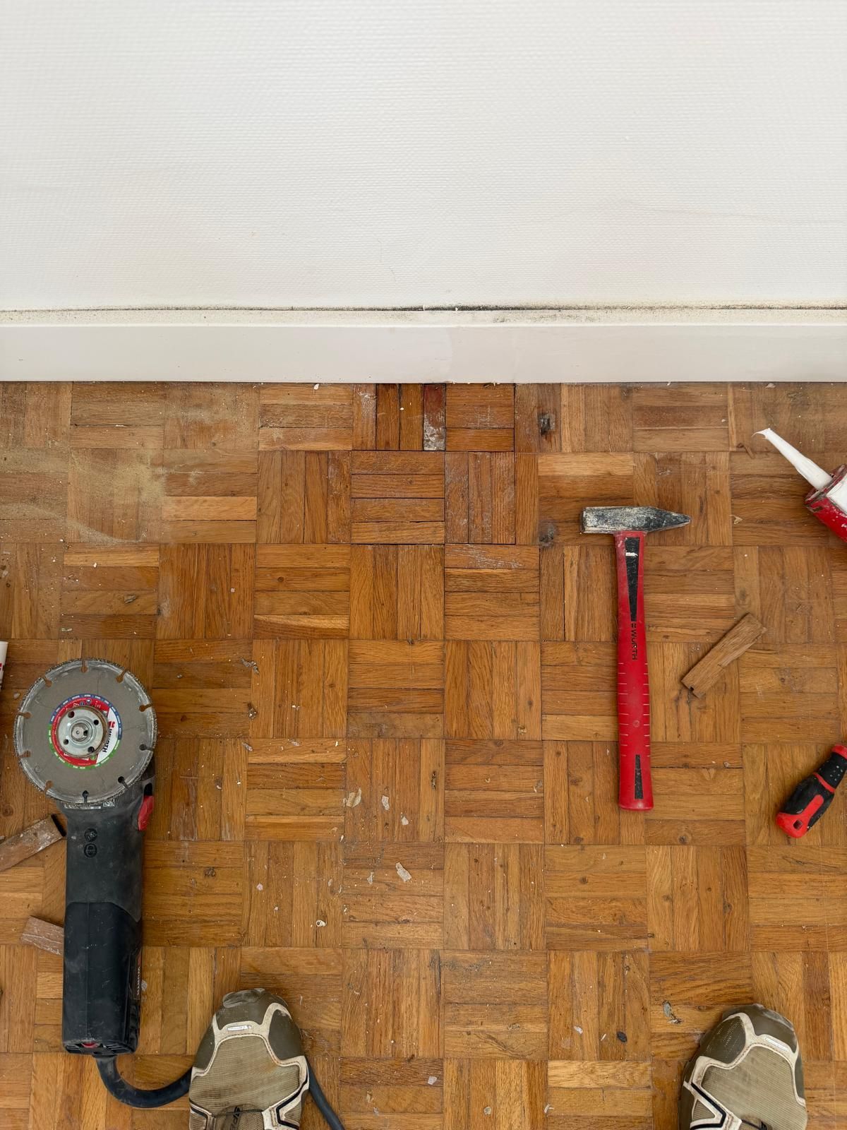 Les pieds d'une personne sur un parquet, près d'un mur blanc, avec des outils : meuleuse d'angle, marteau, pistolet à calfeutrer.