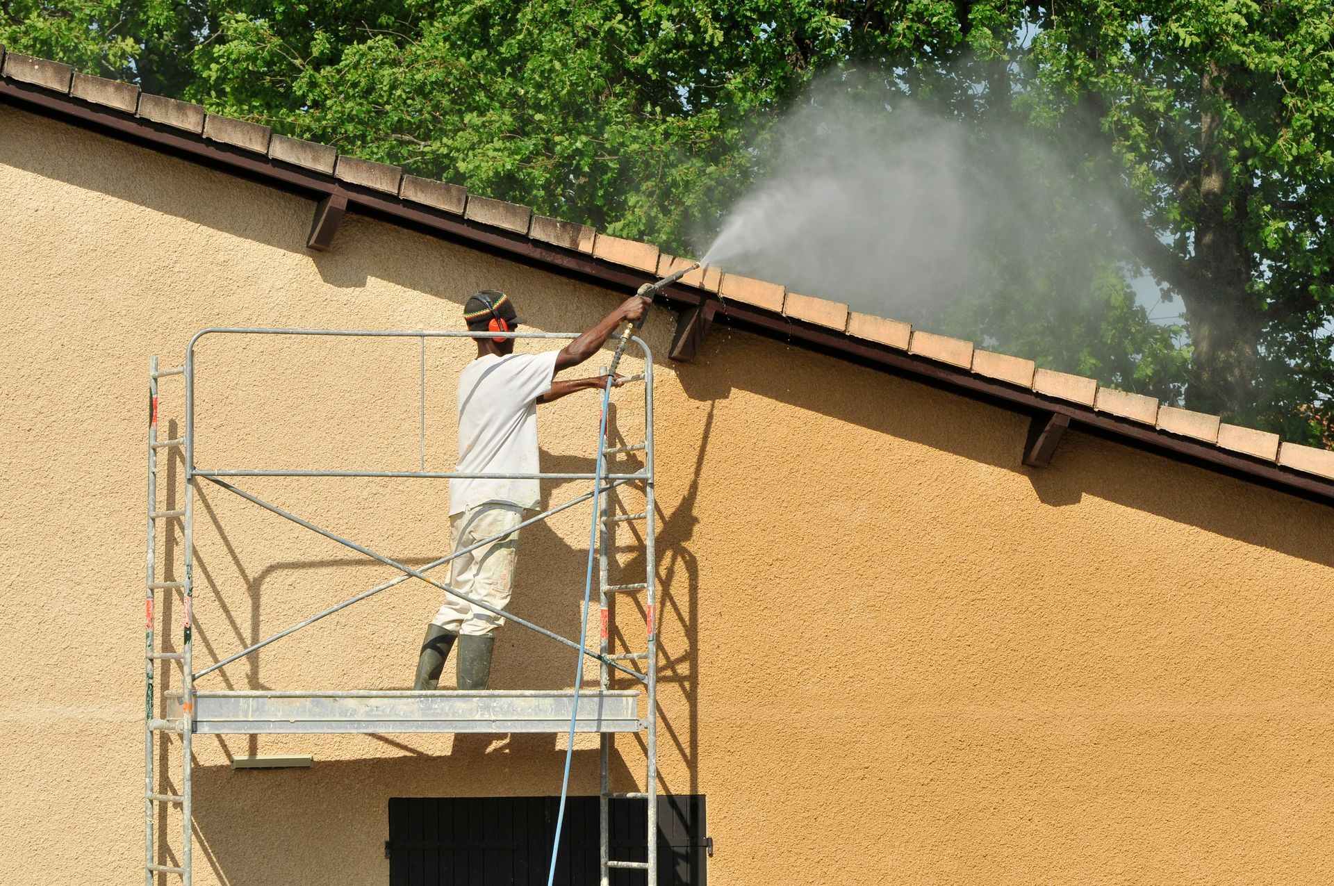 Un homme, juché sur un échafaudage, nettoie la façade d'un bâtiment au nettoyeur haute pression. L'eau jaillit de la buse ; le bâtiment est beige.