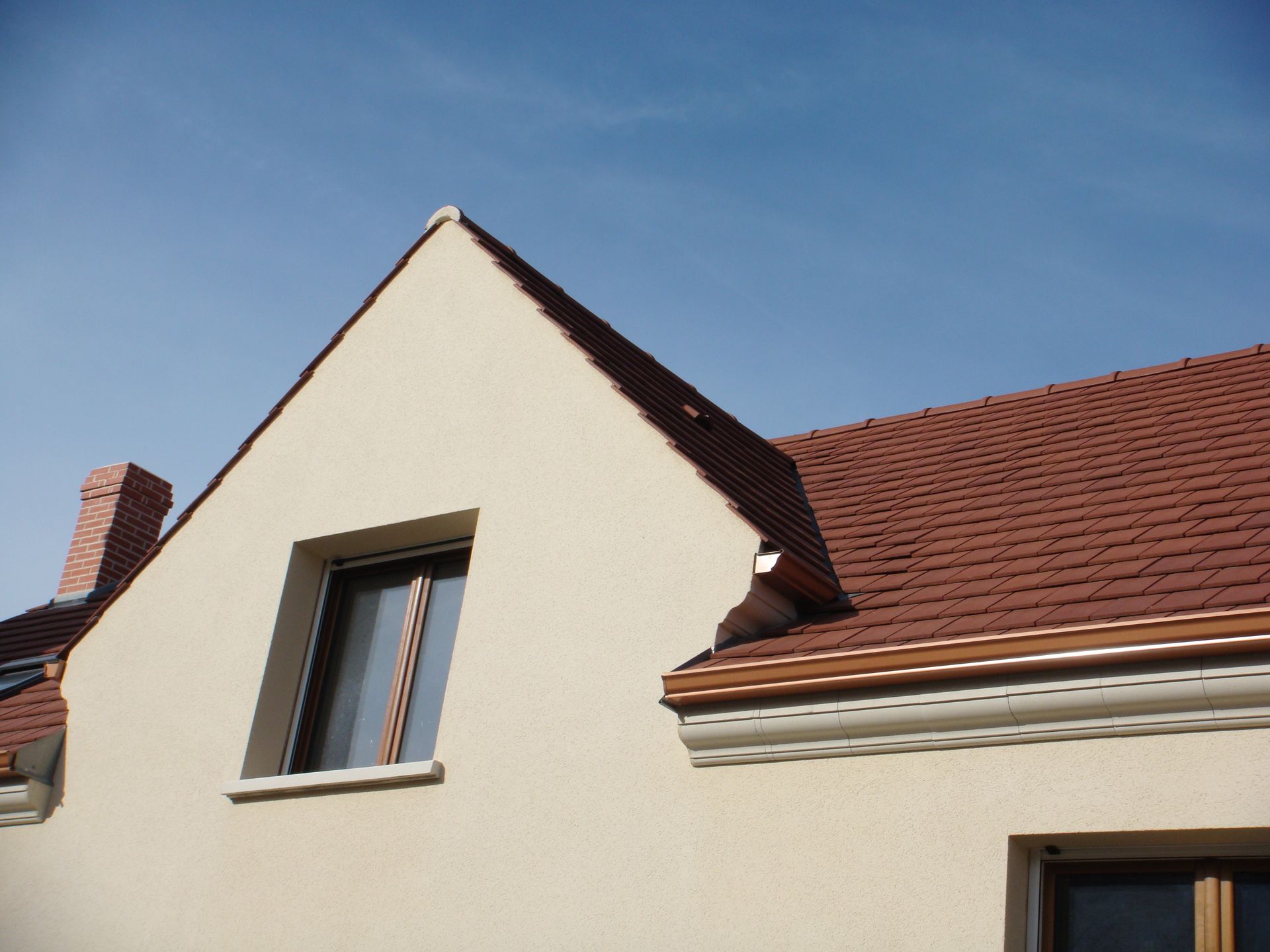 Maison beige avec un toit de tuiles rouges, une fenêtre et un ciel bleu clair.