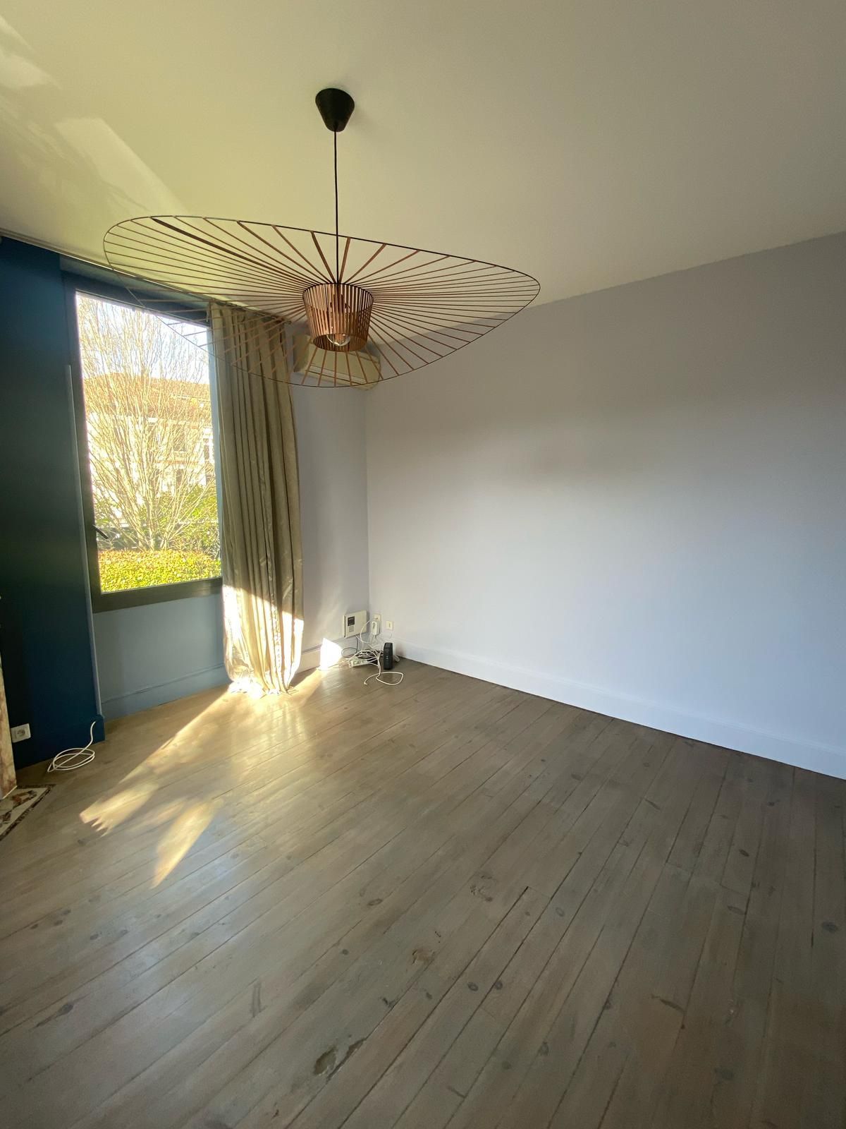 Pièce vide avec parquet, fenêtre avec rideau, luminaire en cuivre et noir, et murs blancs.