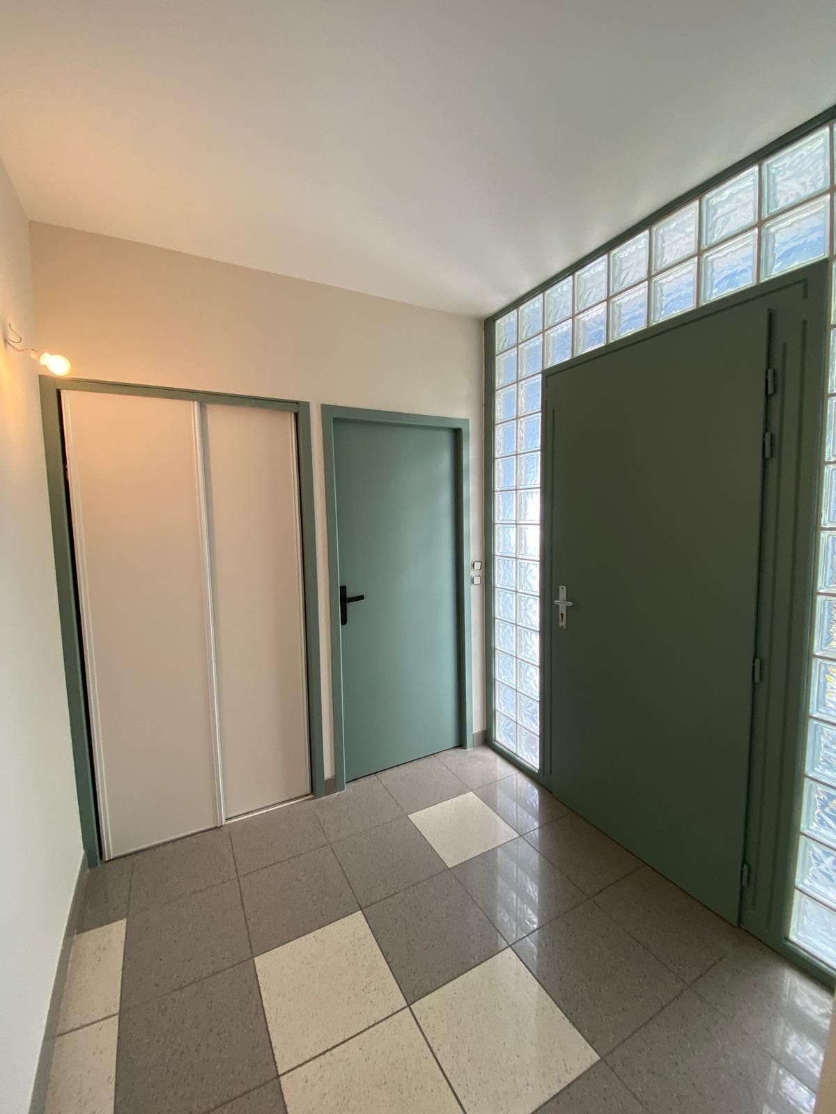 Couloir avec trois portes : portes pliantes blanches, porte verte et porte verte à côté d’un mur en pavés de verre.