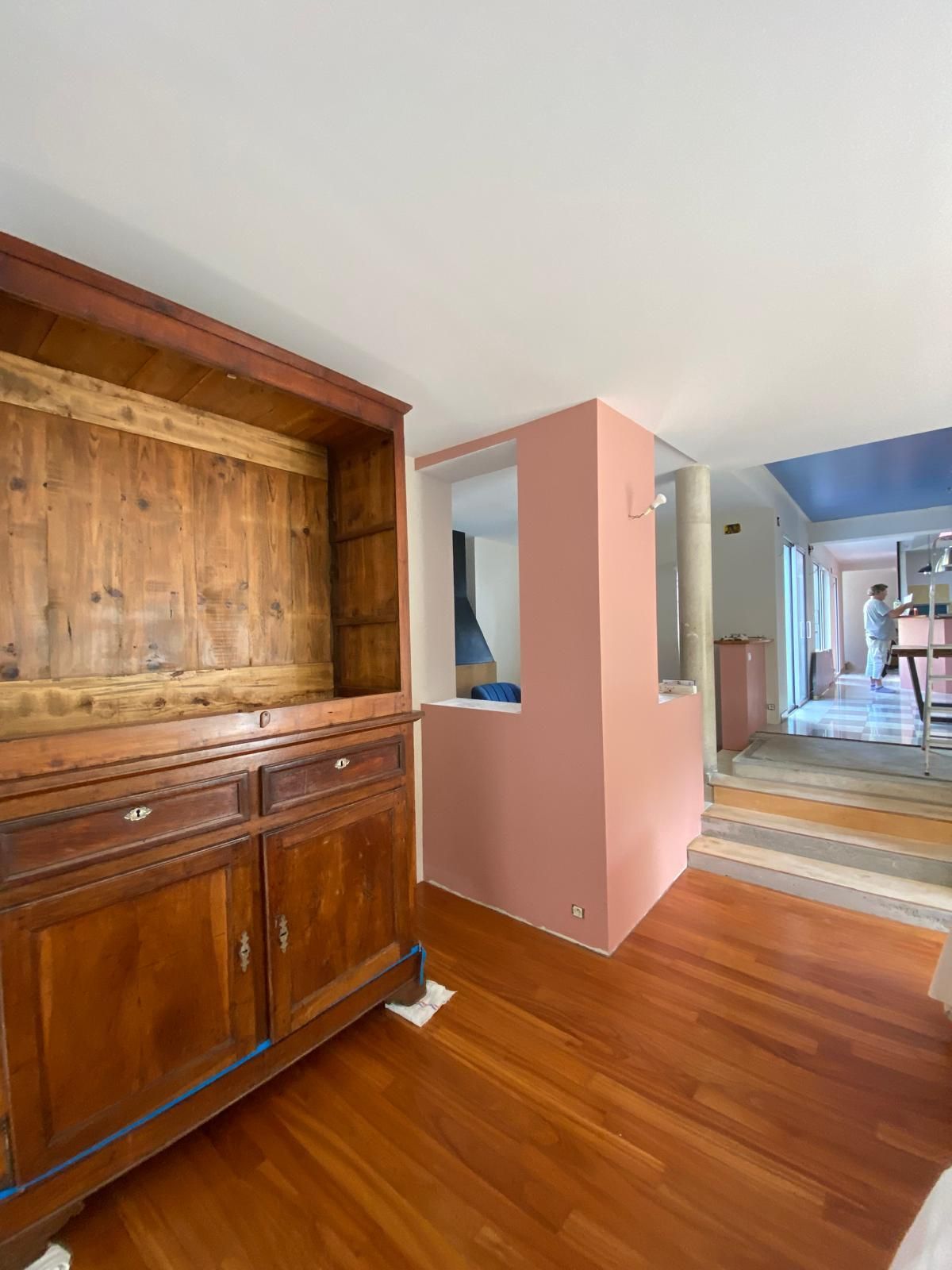Meuble en bois et mur rose dans une pièce au parquet. Une personne marche au loin.