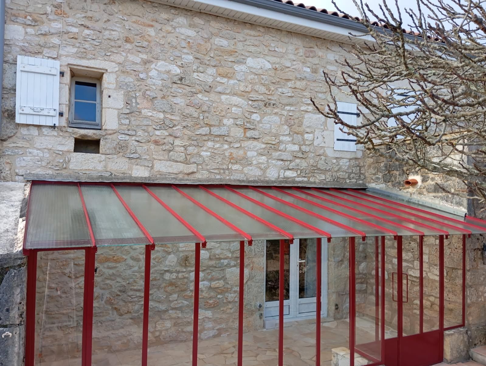 Bâtiment en pierre avec un auvent à cadre rouge au-dessus de la porte. Volets blancs, ciel bleu et arbre sans feuilles visibles.