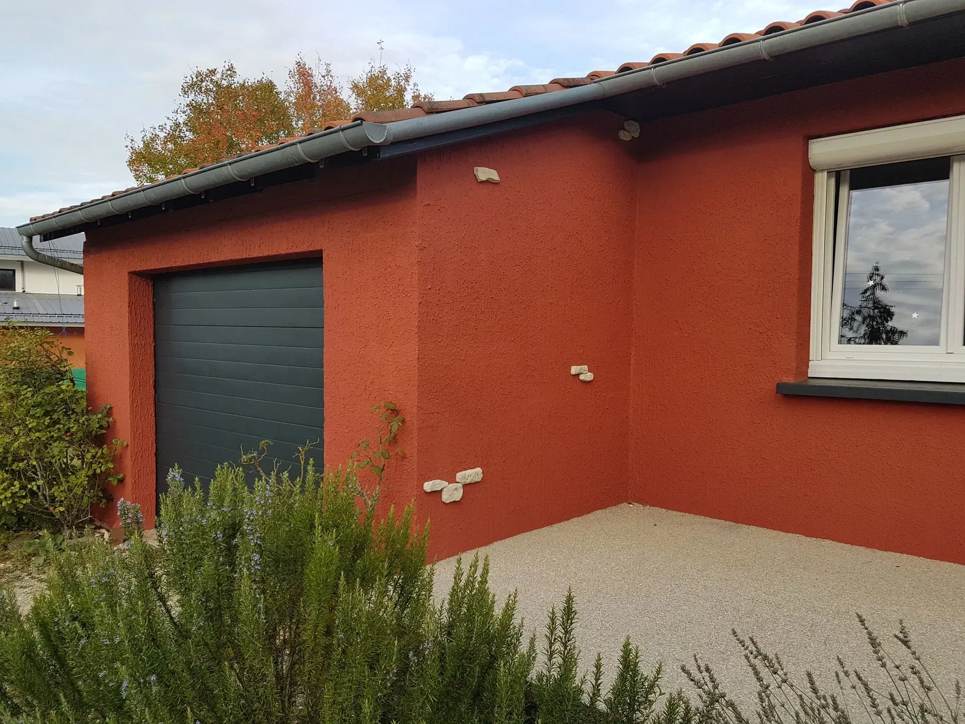 Maison en stuc rouge avec porte de garage grise, fenêtre blanche et terrasse en gravier.