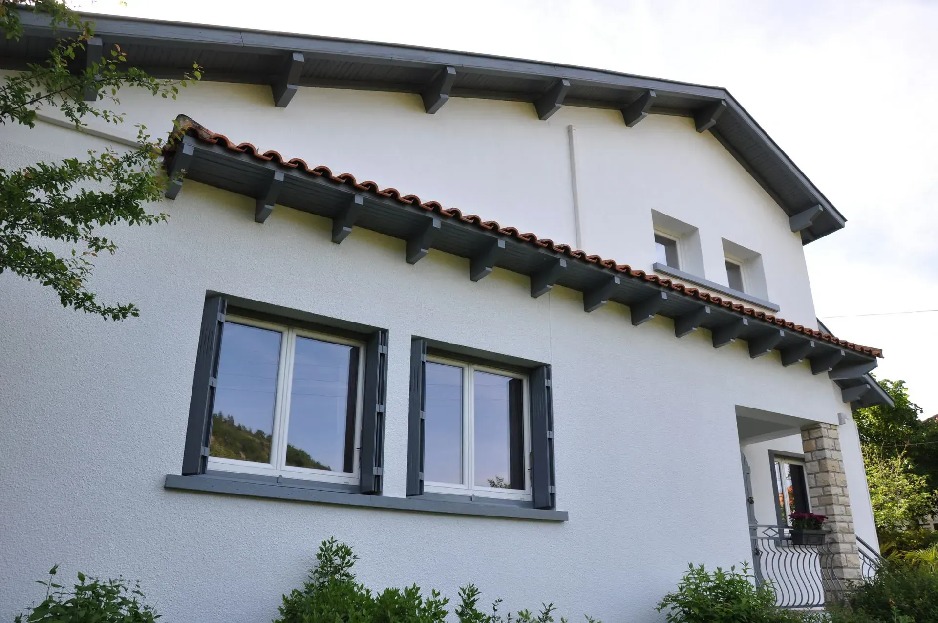 Maison blanche au toit de tuiles rouges, aux boiseries grises et dotée de deux fenêtres en façade.