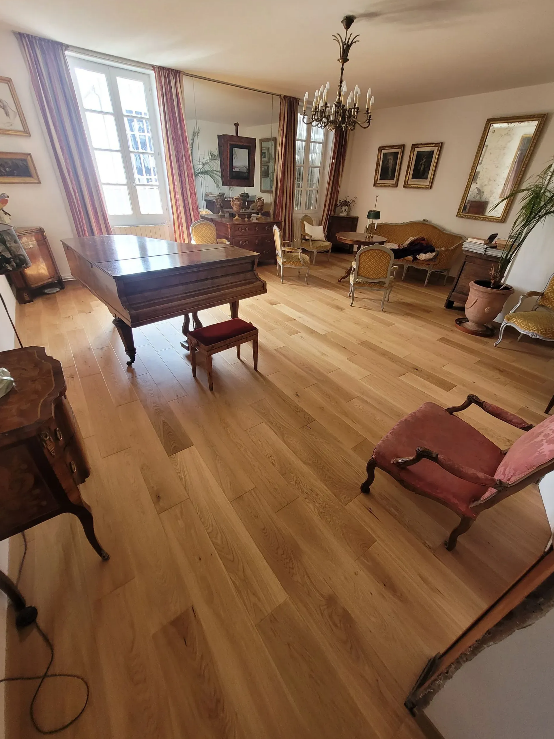 Chambre élégante avec piano, meubles anciens, parquet, lustre et hautes fenêtres.