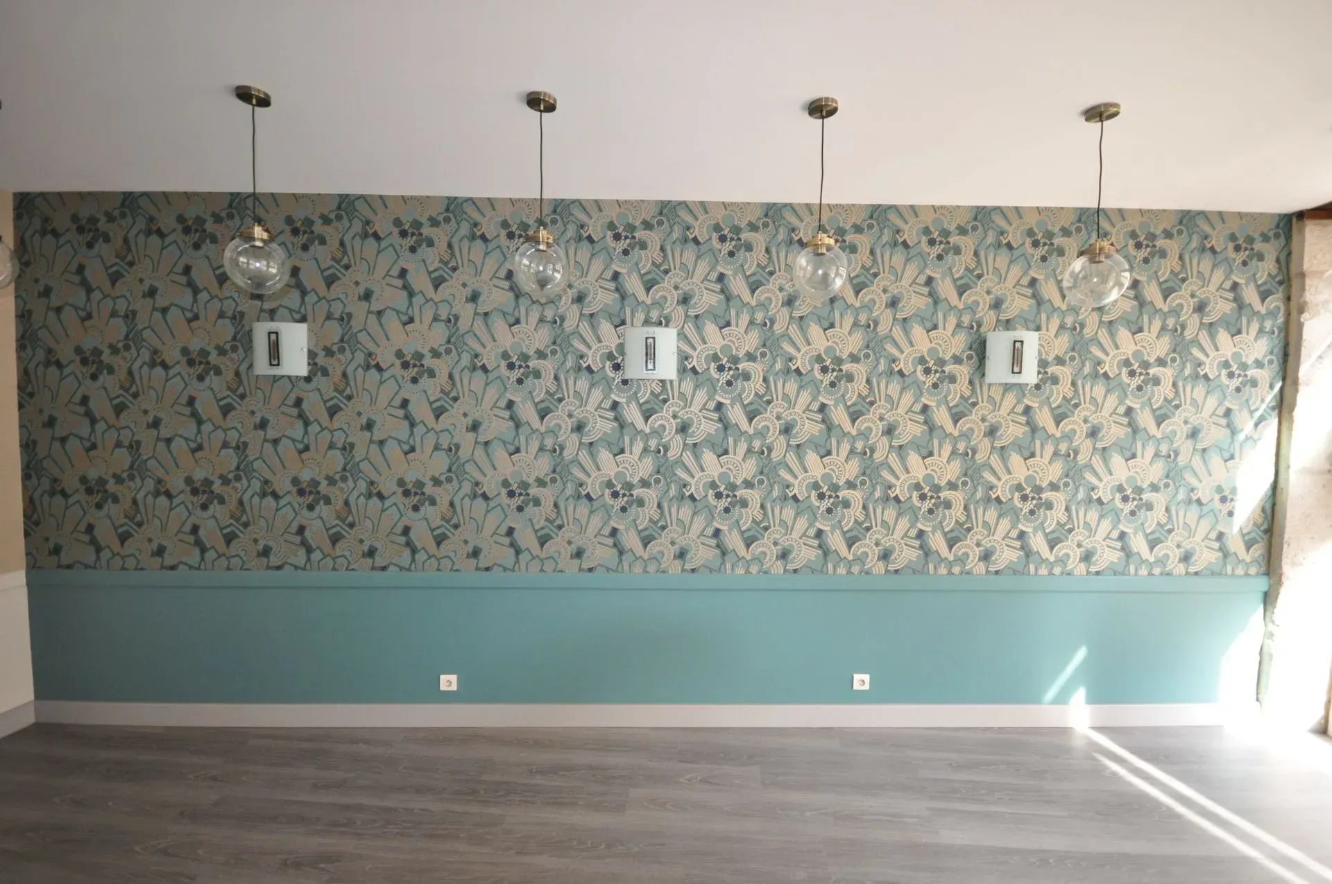 Pièce avec papier peint à motifs turquoise et or, lambris turquoise et suspensions lumineuses. Sol en bois.