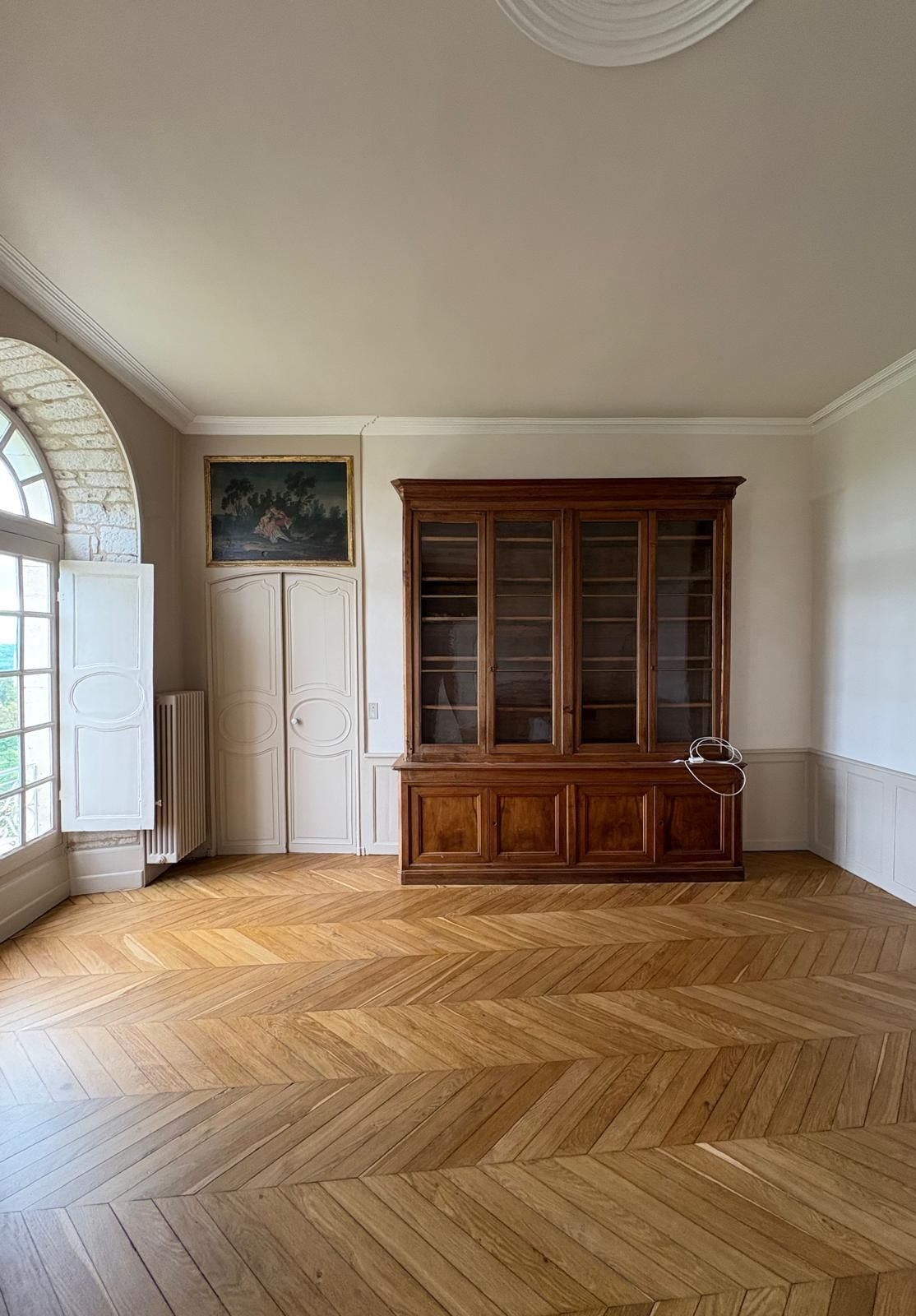 Pièce vide avec parquet, grande armoire en bois, haute fenêtre et œuvres d'art.