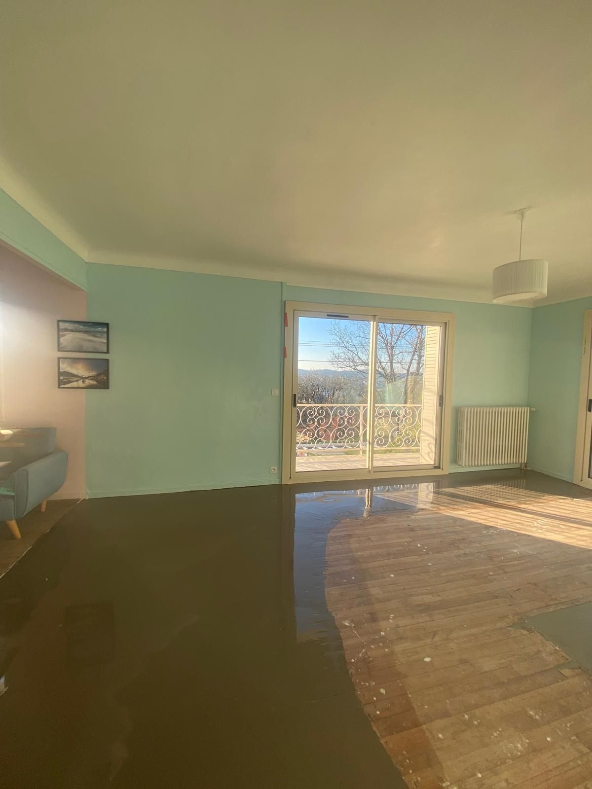 Intérieur d'une pièce aux murs bleu clair et au sol abîmé ; une porte coulissante en verre donne sur un balcon.