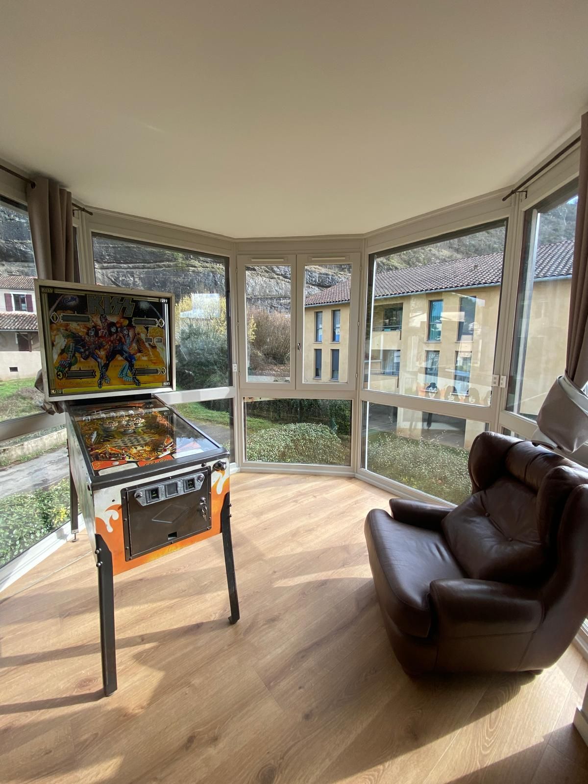 Machine à flipper dans une véranda avec fauteuil et vue panoramique sur l'extérieur.