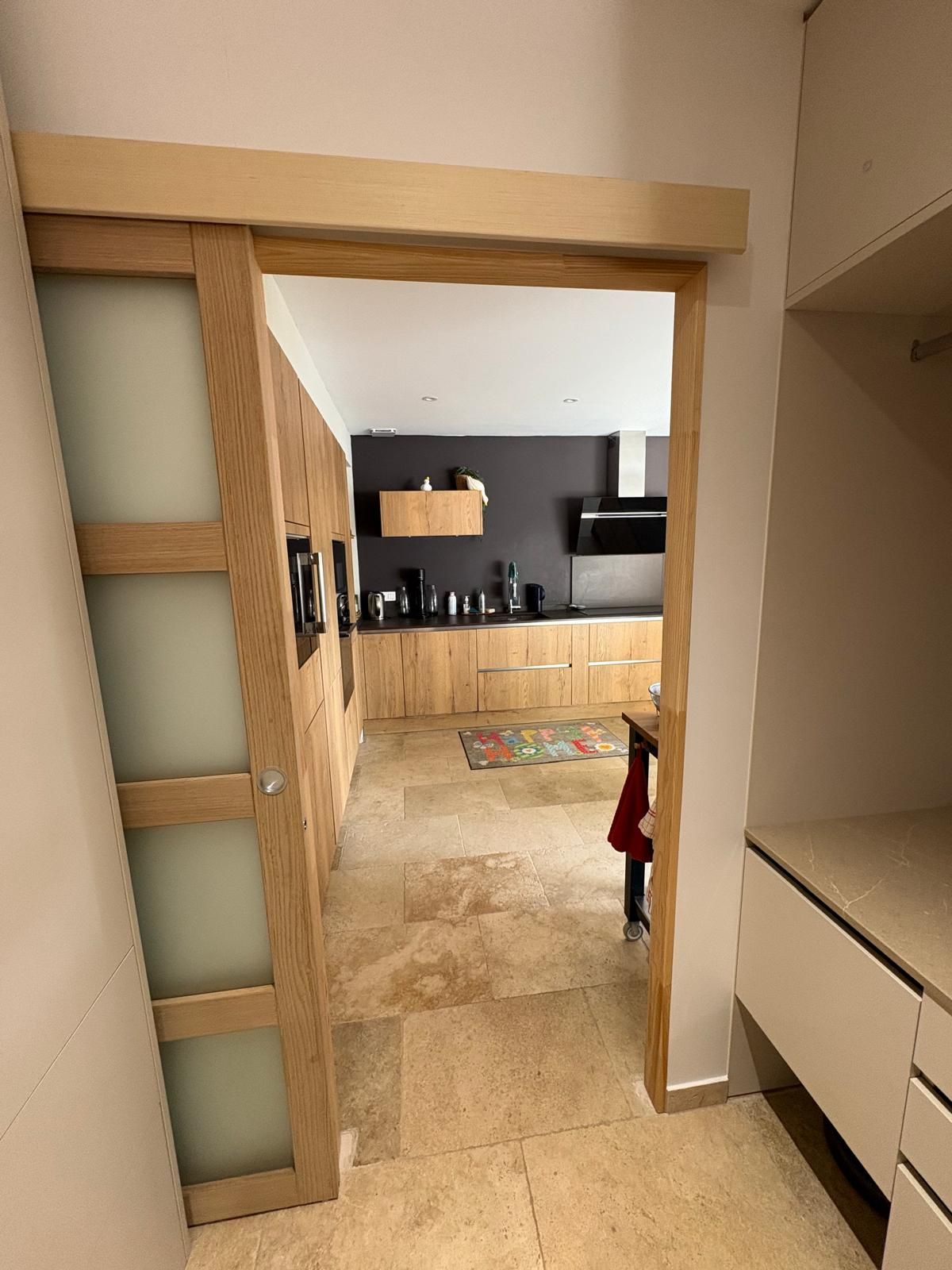 Une porte coulissante ouvre sur une cuisine avec des armoires en bois clair, un sol en pierre et un mur sombre avec une hotte aspirante.