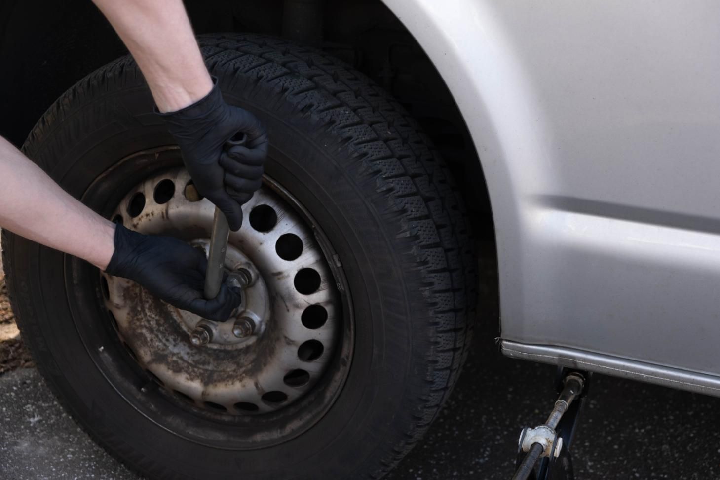 Unas manos enguantadas quitan una rueda de un coche con una llave de tuercas junto a un vehículo plateado.