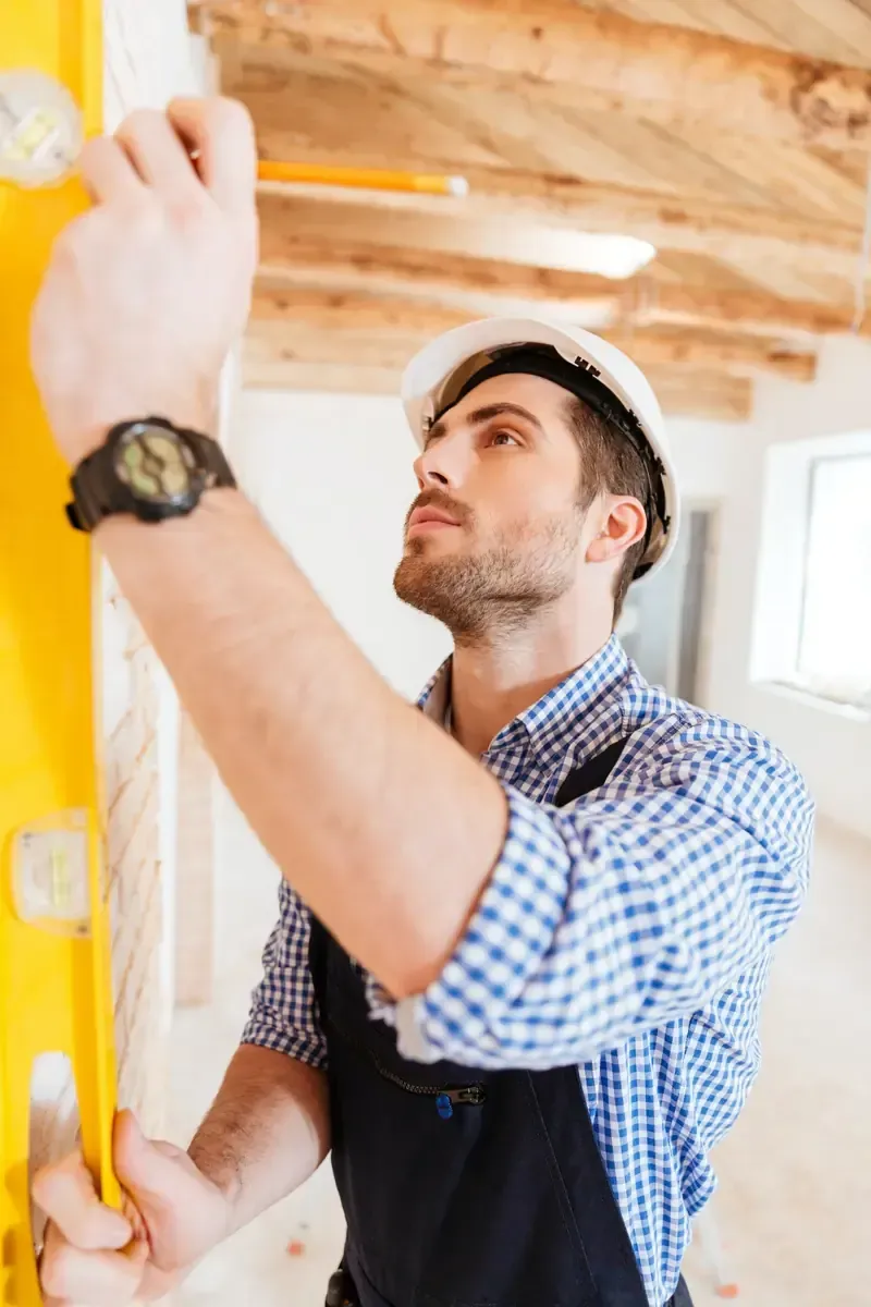 Obrero de la construcción usando un nivel. Con casco y camisa azul a cuadros, dentro de un edificio.