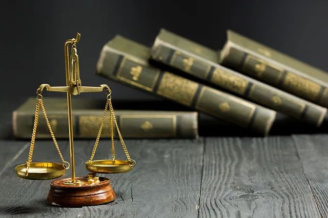 Dos abogados trajeados sentados a la mesa, uno escribiendo y el otro con un libro. Se ven la balanza de la justicia y el mazo.