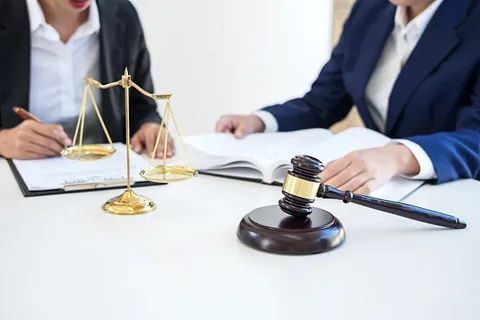 Dos abogados trajeados sentados a la mesa, uno escribiendo y el otro con un libro. Se ven la balanza de la justicia y el mazo.
