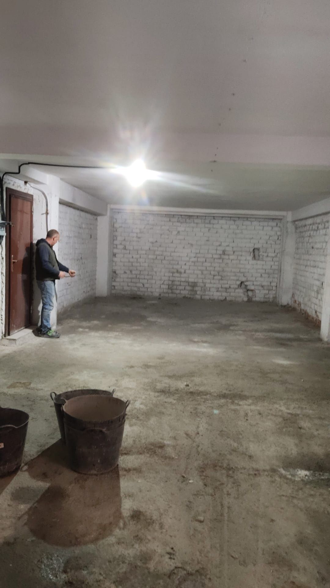 Hombre en un garaje, señalando; piso de concreto, paredes de ladrillo, cubos.