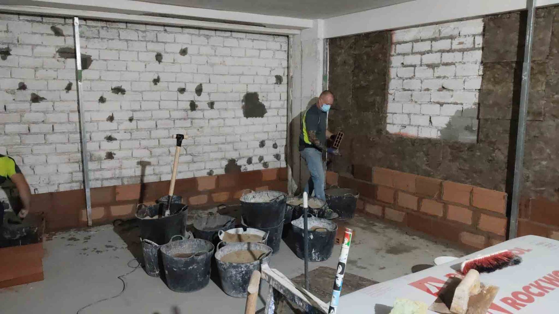 Obreros de la construcción dentro de una habitación, paredes de ladrillo, baldes de material.