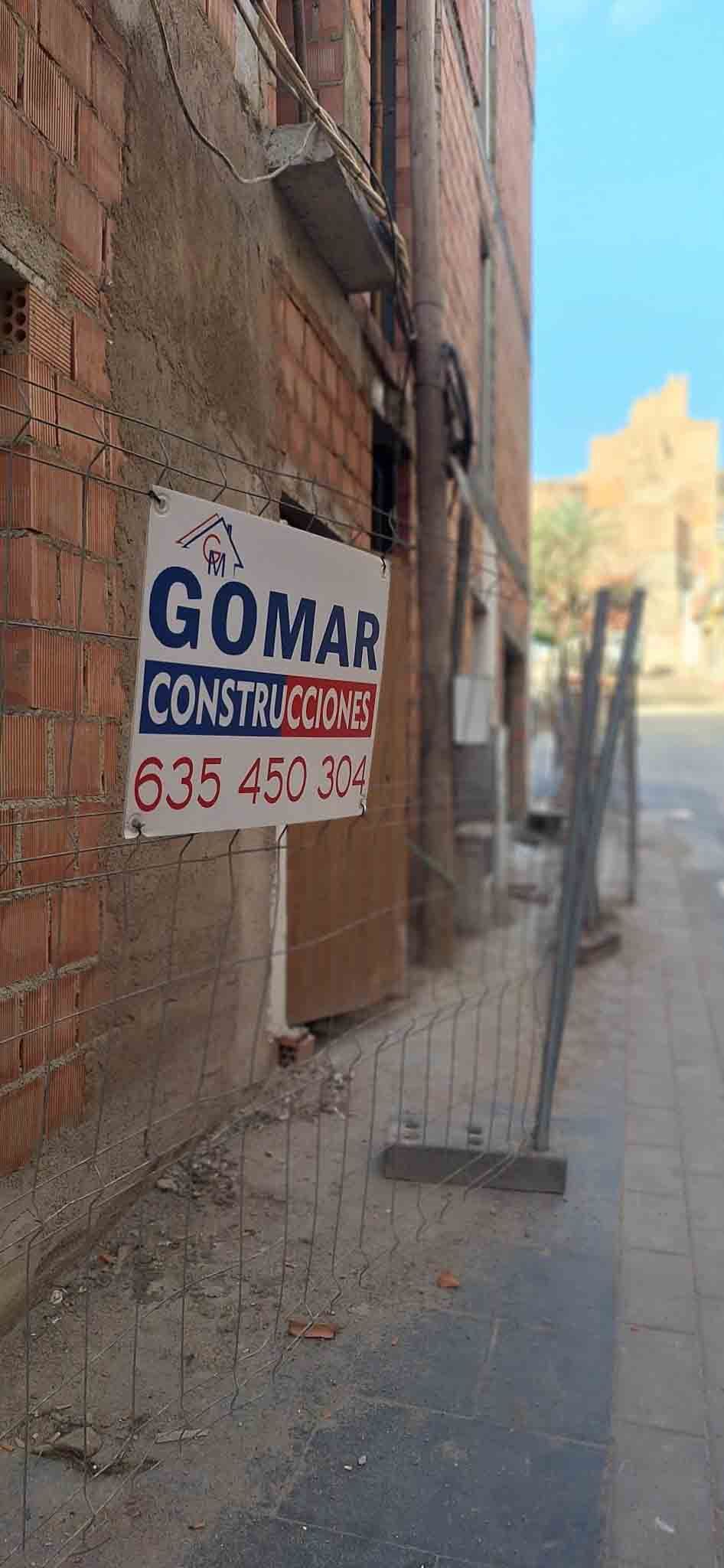 Un edificio con cartel de Gomar Construcciones, con pared de ladrillo y acera, en un día soleado.