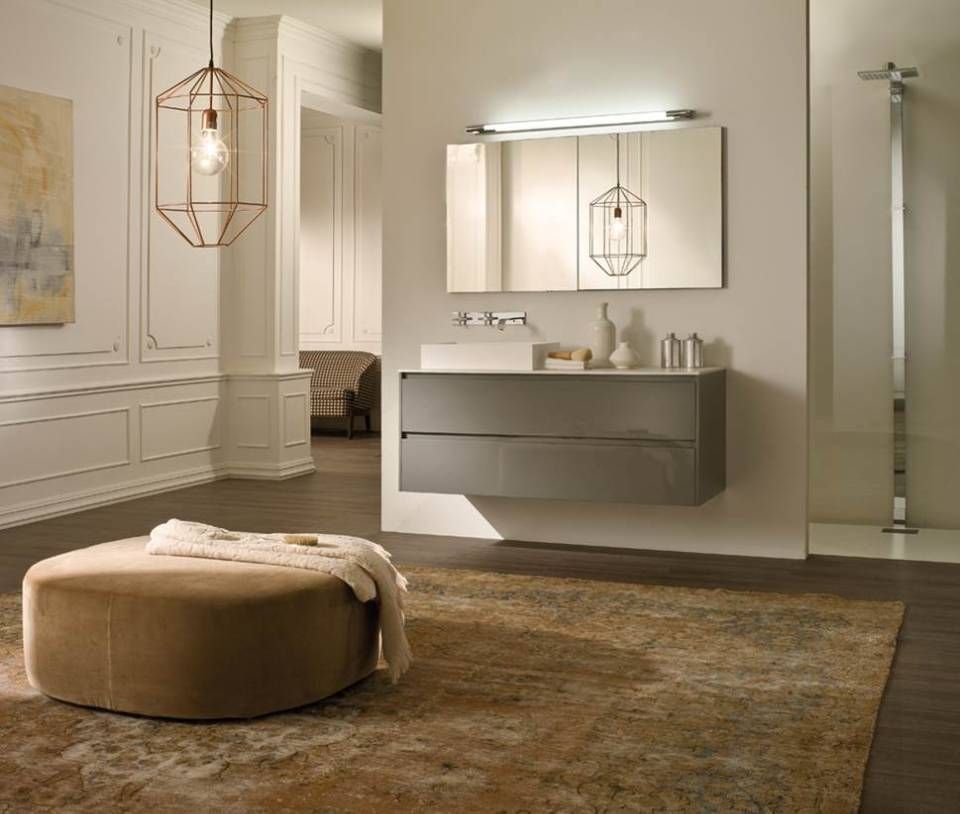 Ein modernes Badezimmer mit einem schwebenden grauen Waschtisch, einem Wandspiegel, einer geometrischen Pendelleuchte und einem hellbraunen Hocker auf einem Teppich.