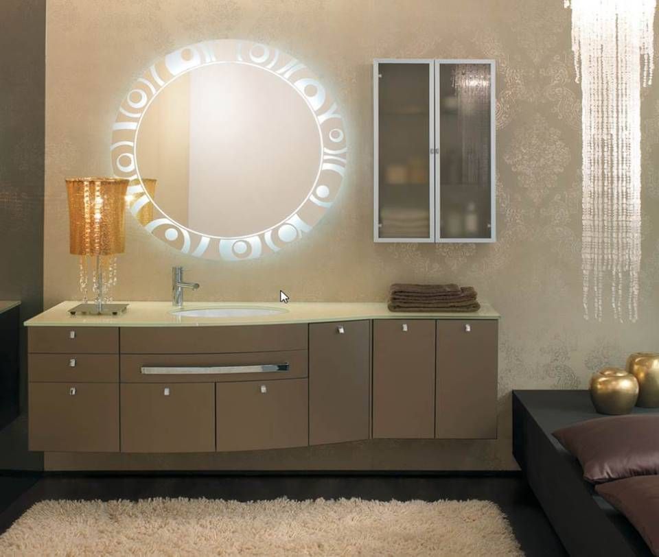 Ein moderner Badezimmerwaschtisch mit beleuchtetem runden Spiegel, hellbraunen Schränken, einem Wandschrank mit Glastür und beigefarbener Dekoration.