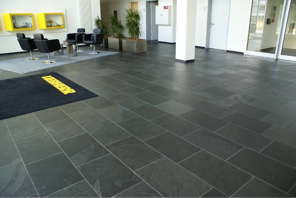 Eine moderne Bürolobby mit dunkelgrauen Steinfliesen, schwarzen Möbeln und hohen Topfpflanzen.