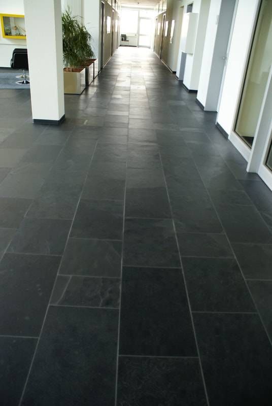 Ein langer, heller Flur mit dunkelgrauen, rechteckigen Bodenfliesen, weissen Säulen und Topfpflanzen an einer Seite.