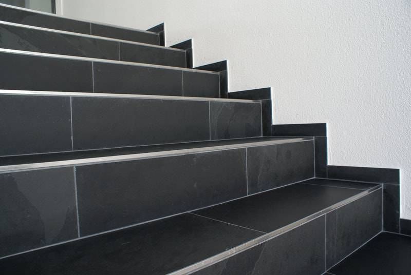 Eine Treppe aus dunklen, schieferartigen Fliesen mit dünnen Metallkanten vor einer schlichten weissen Wand.