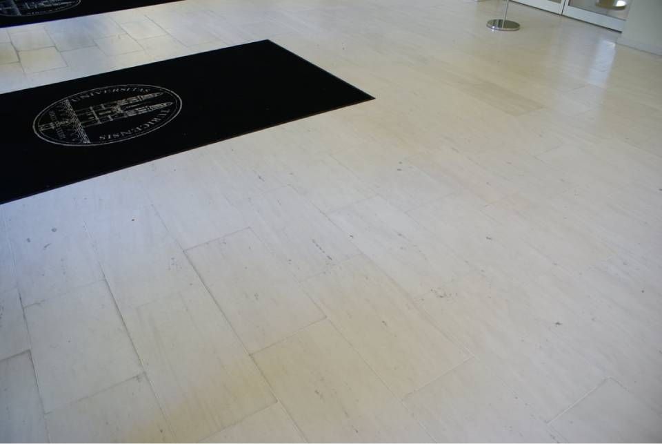 Eine schwarze Fußmatte mit einem runden Logo liegt auf einem hellen Steinfliesenboden in einem Innenraum.