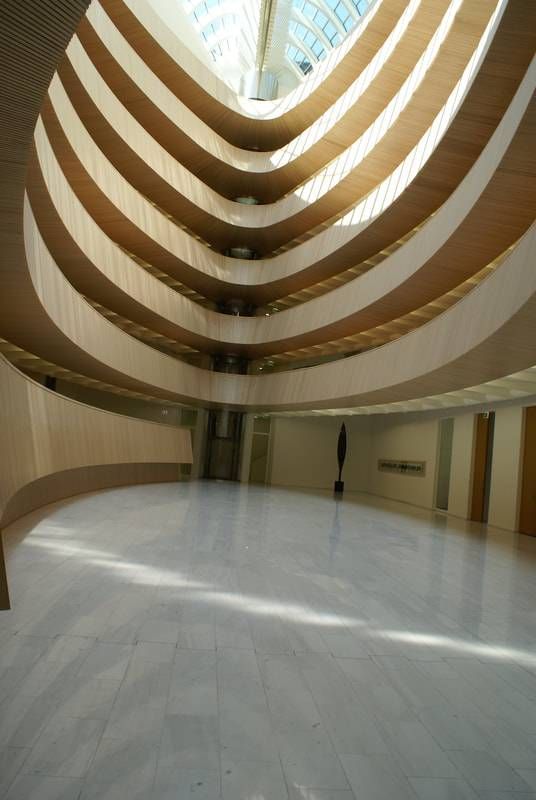 Eine weitläufige, sonnendurchflutete Lobby mit einem hohen, geschwungenen Atrium und mehrstufigen, hellen Holzbalkonen.