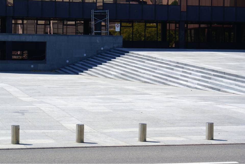 Ein weitläufiger, gepflasterter öffentlicher Platz mit einer markanten, breiten, gestaffelten Treppe, die zu einer modernen Gebäudefassade hinaufführt.
