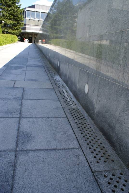 Ein graues Steingebäude mit einem gepflasterten Weg und einem linearen Metall-Entwässerungsgitter, das entlang des Sockels der Mauer verläuft.
