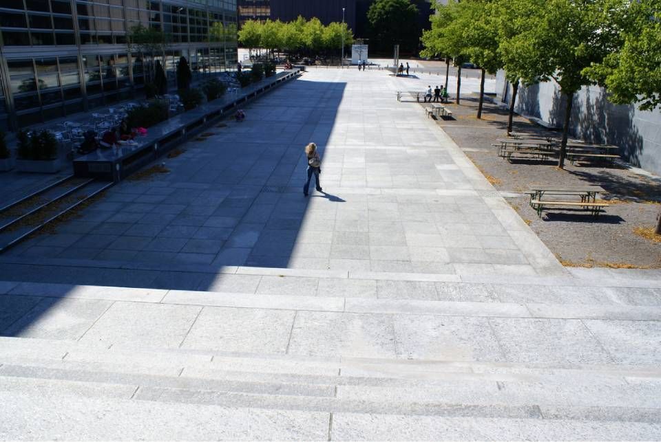 Eine Vogelperspektive auf einen weitläufigen Steinplatz mit einer Treppe im Vordergrund und einer einzelnen Person, die über den offenen Platz geht.