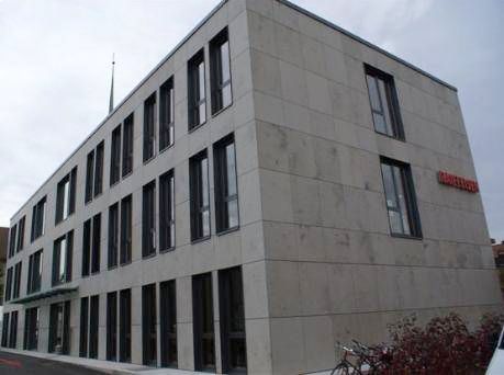 Modernes dreistöckiges graues Betongebäude mit rechteckigen Fenstern und einem roten Schild an der Seite, unter einem bewölkten Himmel.