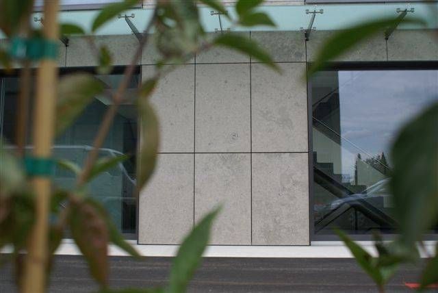 Eine hellgraue, mit Steinpaneelen verkleidete Mauer, eingerahmt von verschwommenen grünen Blättern und Glasfenstern im Freien.