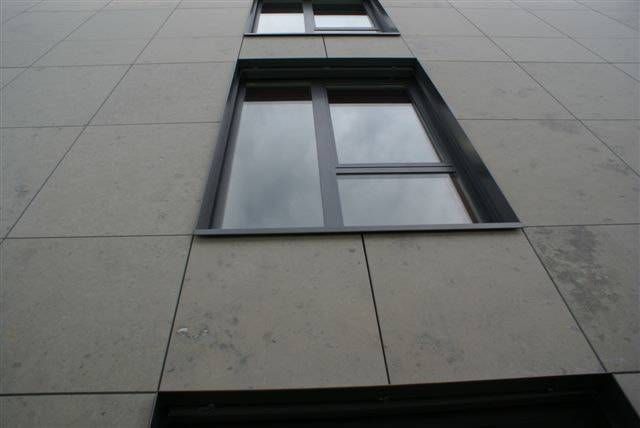 Eine Ansicht aus der Froschperspektive auf zwei rechteckige Fenster in einer grauen, gefliesten Gebäudefassade unter bedecktem Himmel.